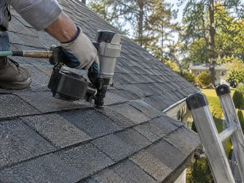 Gloved hand using a nail gun on architectural asphalt shingles, with a ladder at the roof edge